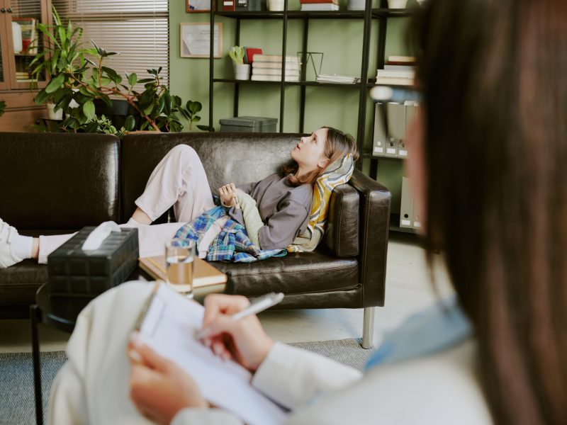 Client laying on couch in cozy office with therapist taking notes on clipboard and bookshelf in background creating inviting atmosphere